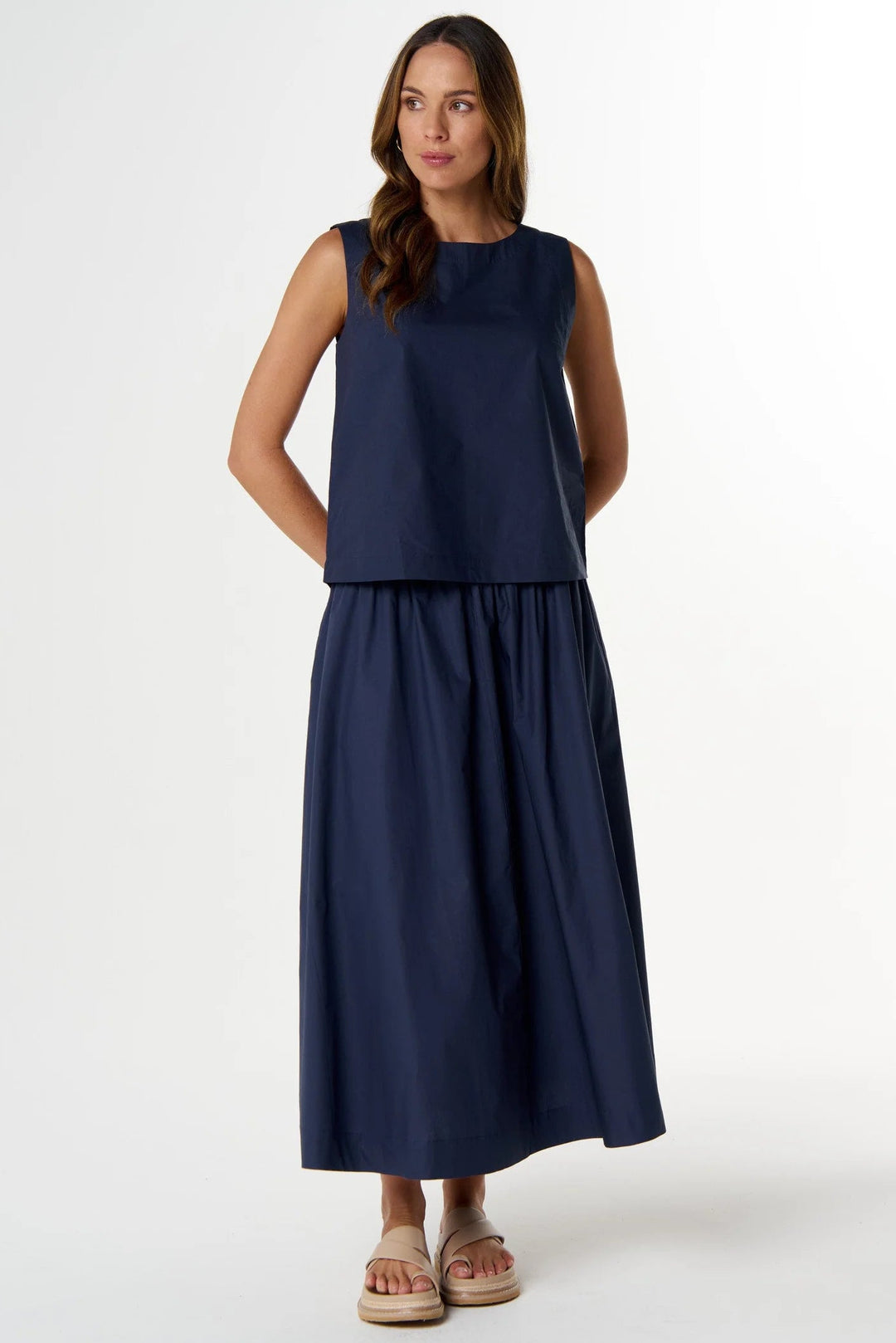 A woman modeling a sleeveless navy blue maxi dress with a cropped top and a flowing skirt, standing against a plain white studio background.