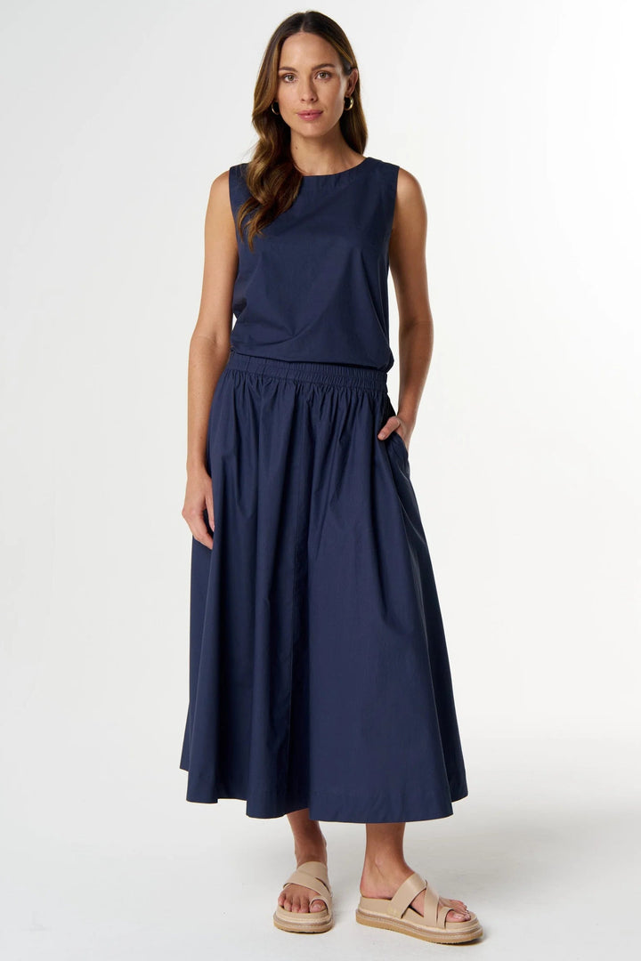 A woman modeling a sleeveless navy blue midi dress with an elastic waist and side pockets, paired with beige espadrille sandals, against a plain white studio background.