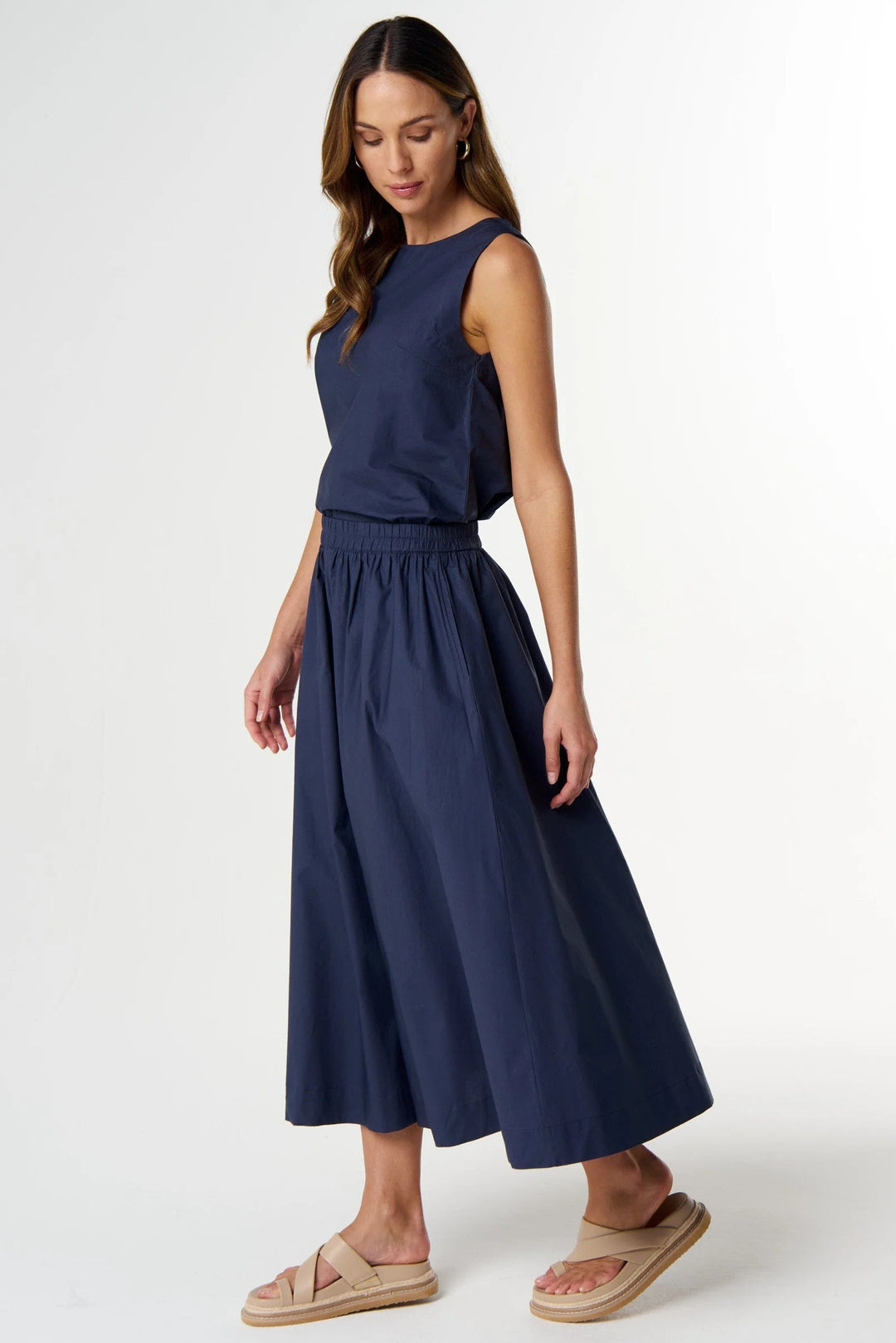 A woman modeling a sleeveless navy blue midi dress with a gathered waist and flared skirt, paired with beige slide sandals, against a plain white studio background.