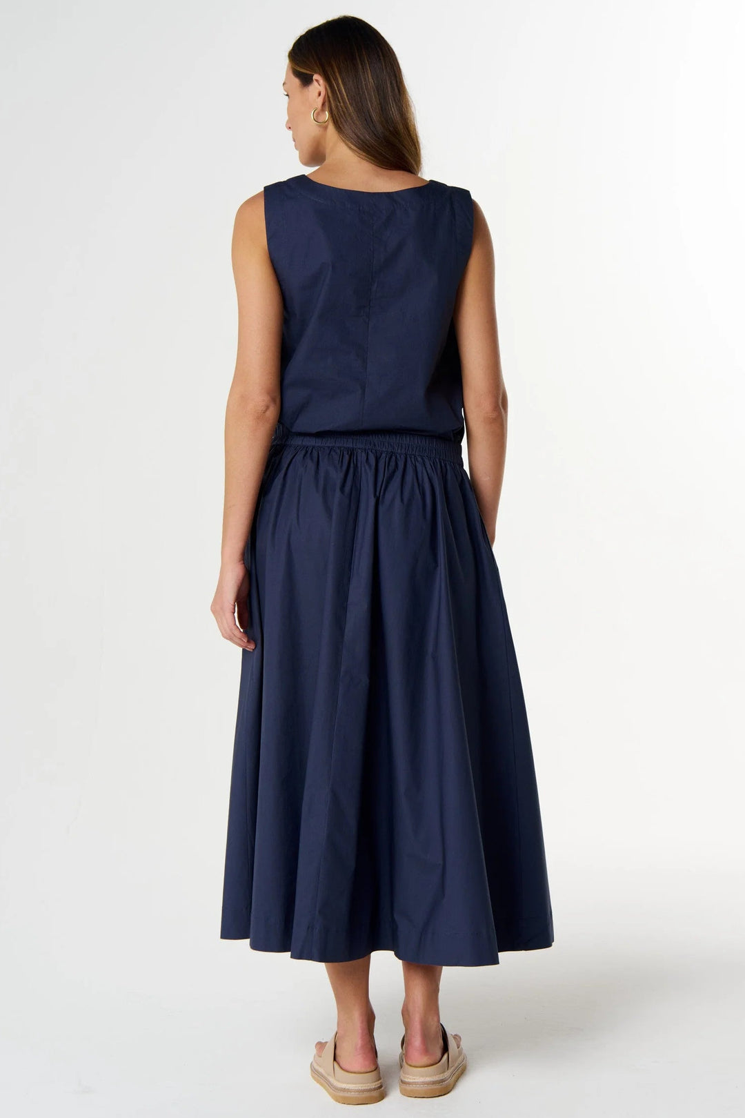 A woman viewed from the back wearing a sleeveless navy midi dress with a gathered waist and espadrille sandals, photographed against a clean white studio background.