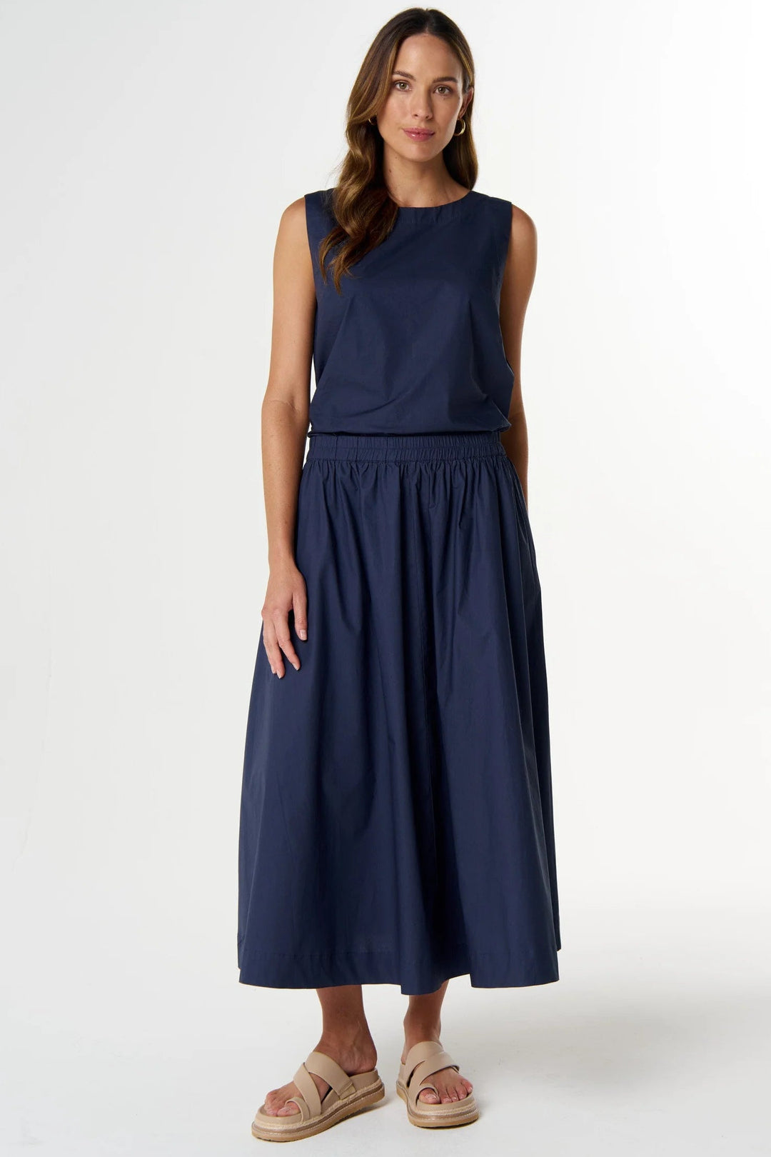A woman modeling a sleeveless navy blue midi dress with a gathered elastic waist, paired with beige espadrille sandals, against a plain white studio background.