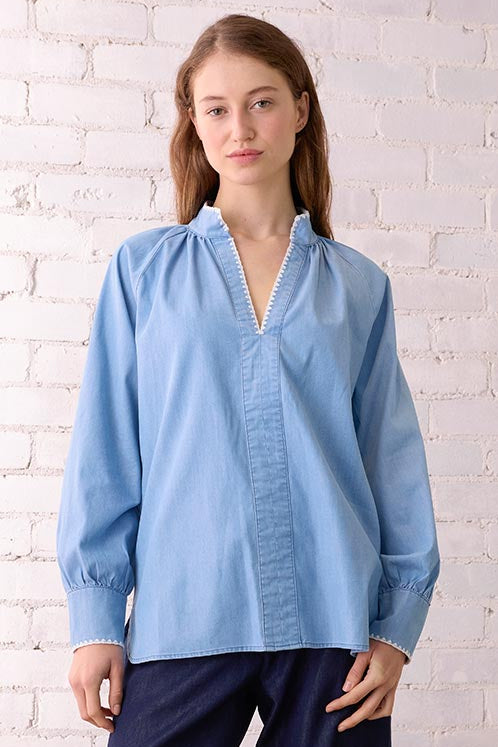 Woman wearing a light blue shirt and dark blue pants standing against a white brick wall.