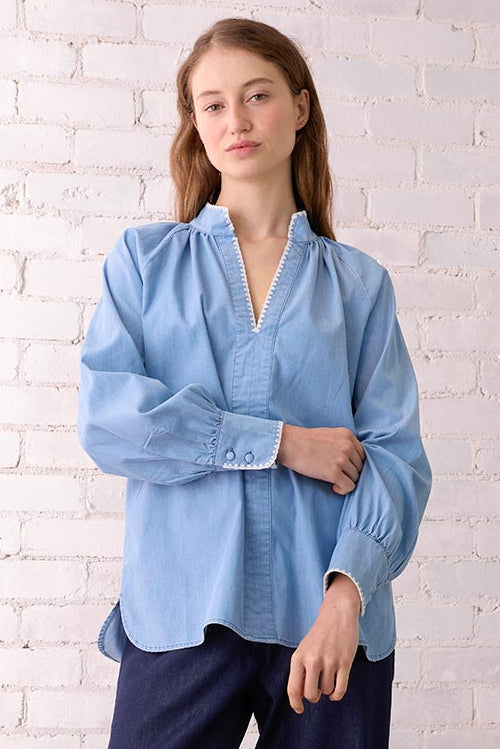 Woman wearing a light blue blouse and dark blue pants standing in a room with a white brick wall and wooden chair.