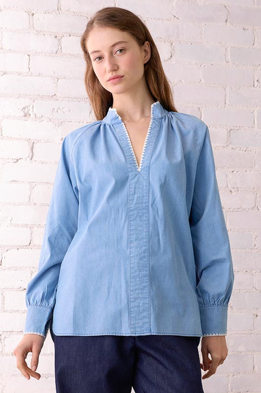Woman wearing a light blue blouse and dark blue pants standing in a room with a white brick wall and wooden chair.