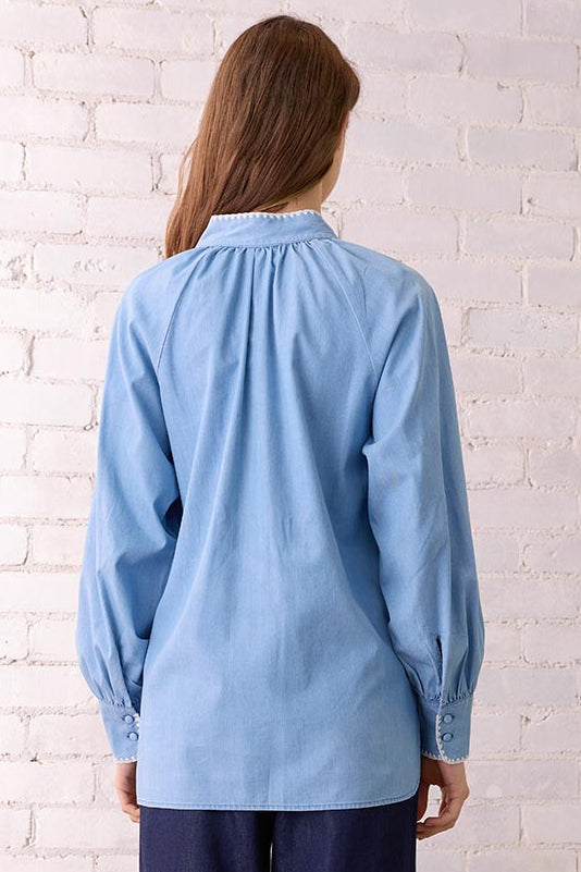 Woman wearing a light blue blouse and dark blue pants standing in a room with a white brick wall and wicker chair.