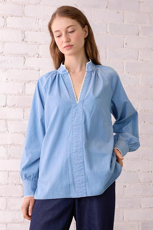 Woman wearing a light blue shirt and dark blue pants standing against a white brick wall.