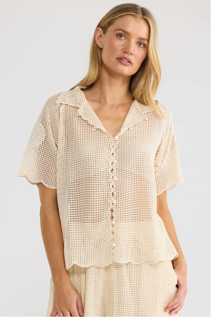 A woman wearing a cream-colored crochet short-sleeve button-up shirt and matching crochet bottoms, photographed against a plain light gray studio background.