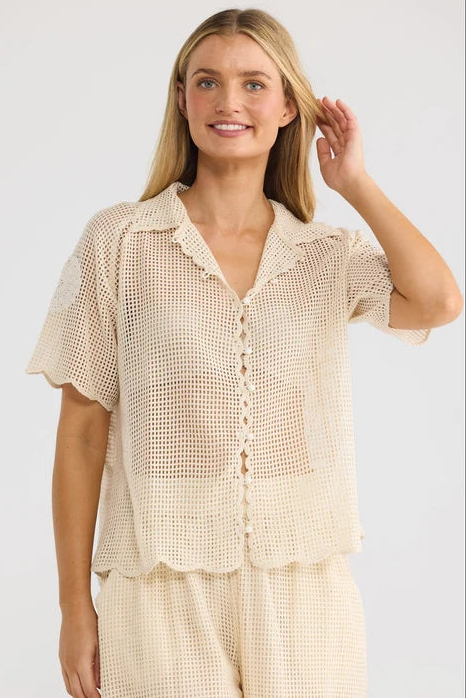 A woman modeling a light beige crochet short-sleeve button-up shirt and matching pants against a plain light gray studio background.