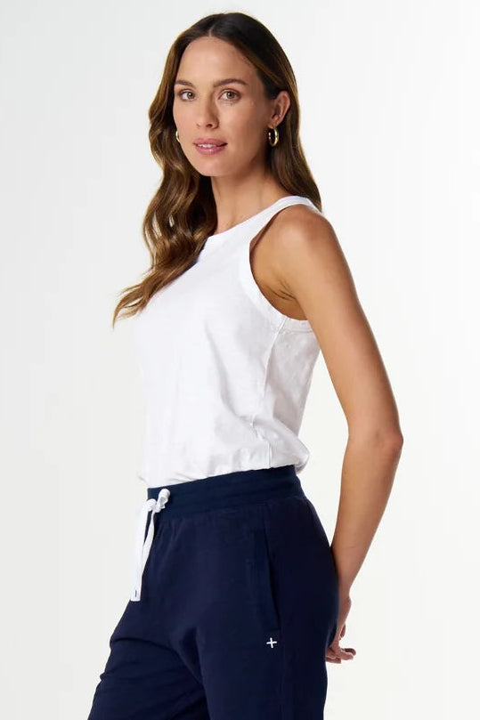 A woman modeling navy blue jogger-style pants, paired with a white sleeveless top and white slide sandals, standing against a clean white studio background.