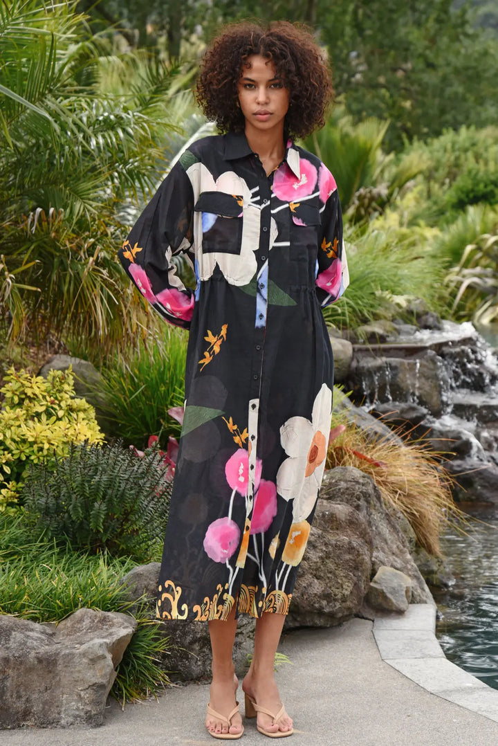 A woman stands outdoors by a garden pond wearing a long black button-front shirt dress with a large floral print in pink, white, green, and gold. The setting includes rocks, tropical plants, and a small waterfall.