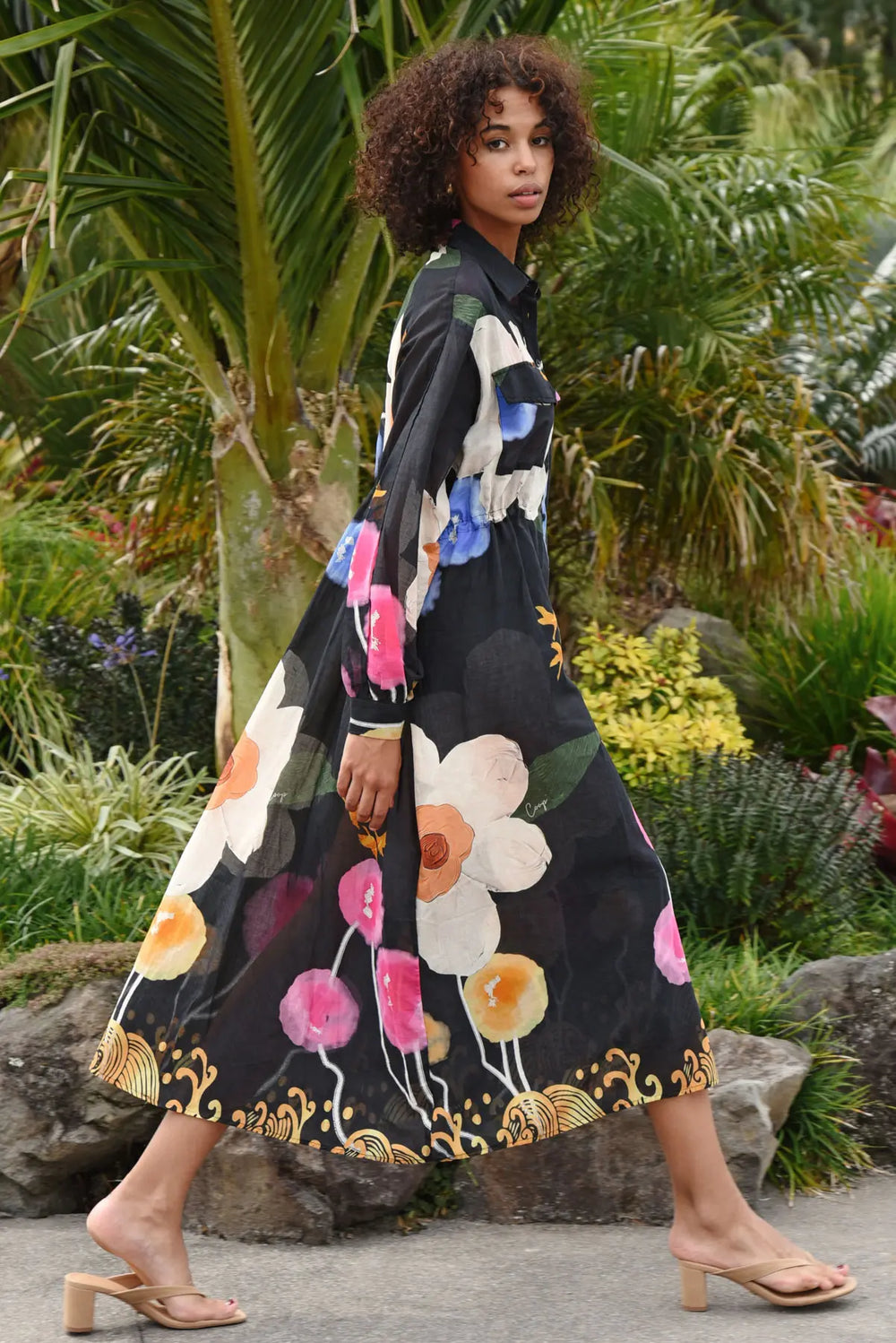 A woman wearing a long-sleeved black midi dress with a colorful floral print and gold decorative trim, walking in a lush tropical garden setting.