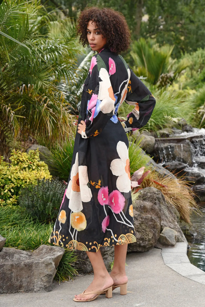 A woman wearing a black midi wrap dress with a vibrant floral print in pink, white, orange, and gold, standing in a lush garden setting.