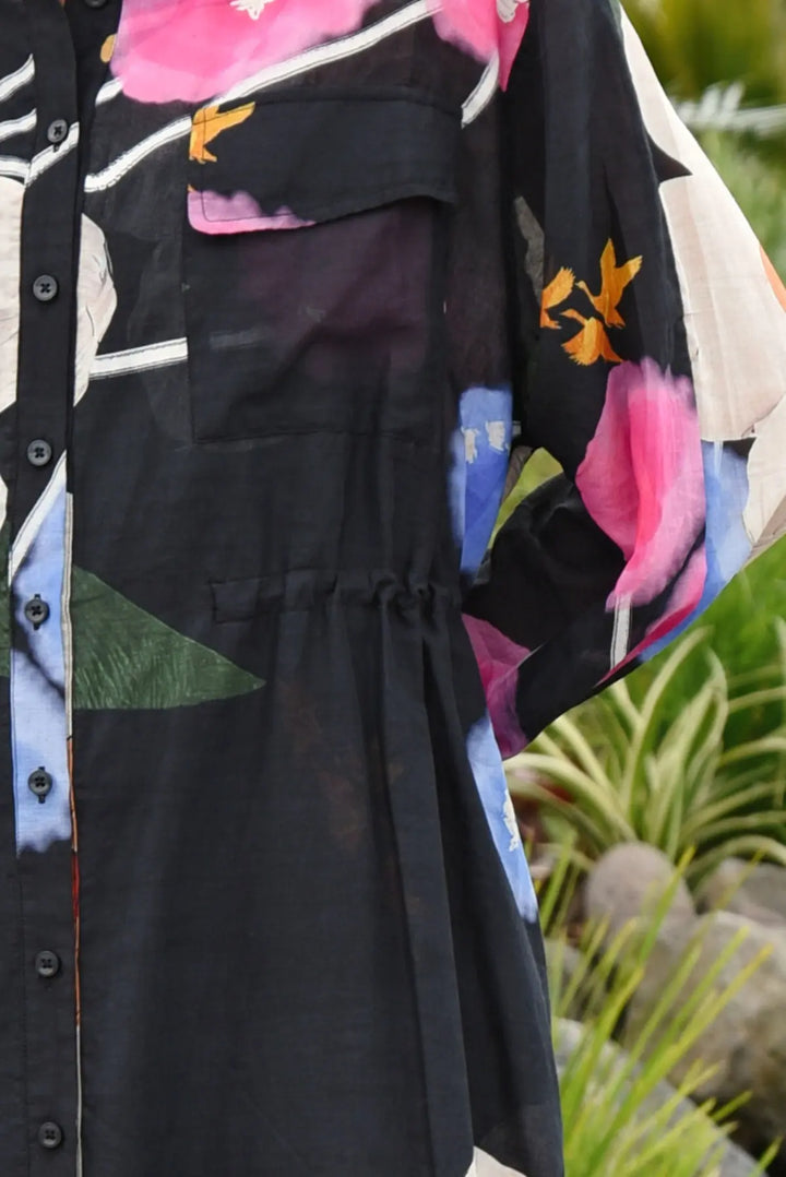 A close-up of a black button-front shirt dress with a vibrant multicolor floral print, featuring pink, blue, green, and orange accents, set against a blurred outdoor garden background.