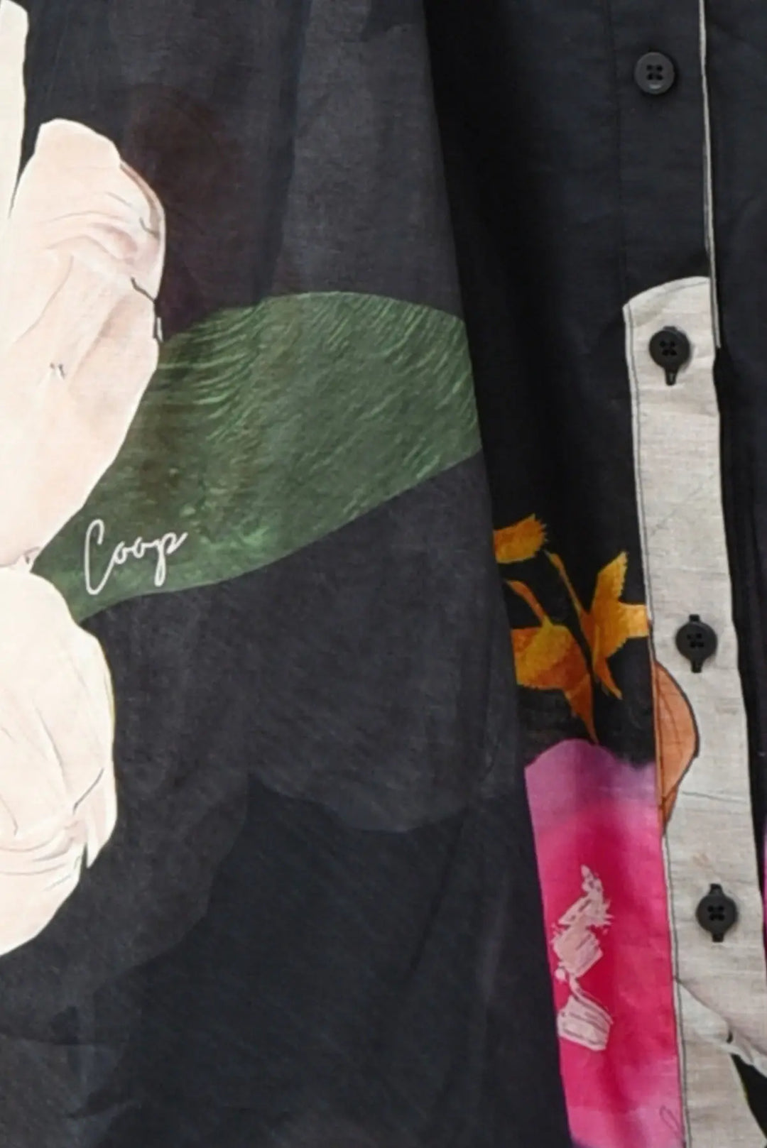 Close-up of a black button-front shirt with a large floral print in pink, green, and white, plus a light gray placket and visible 'Coop' script on the fabric.
