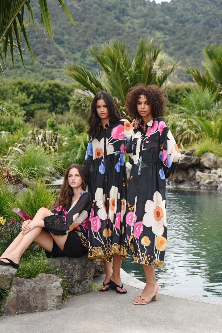 Three women posing outdoors by a pool with lush tropical greenery and mountains in the background. Two women are wearing matching black midi dresses with a vibrant multicolor floral print, and one woman is seated on a rock wearing a black dress.