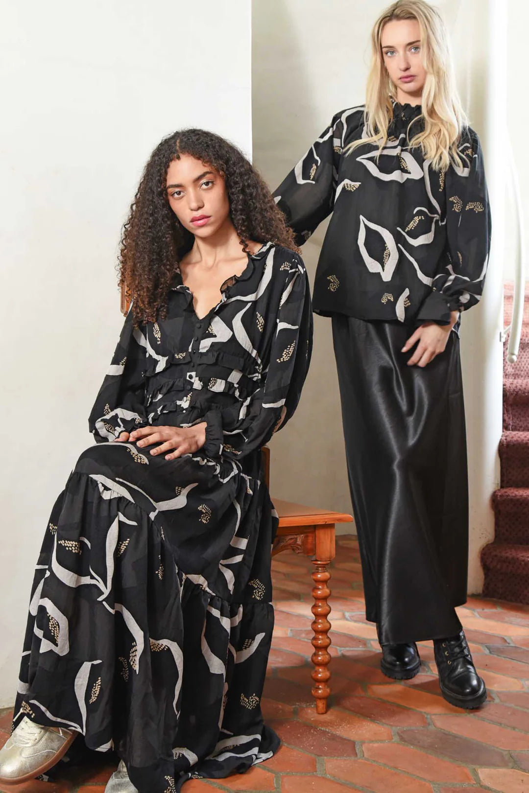 Two women wearing black and white patterned outfits in a room with a wooden table and chair.
