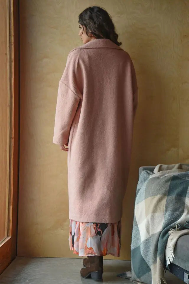 Person wearing a long pink coat standing in a room with a textured wall and a blanket on a chair.