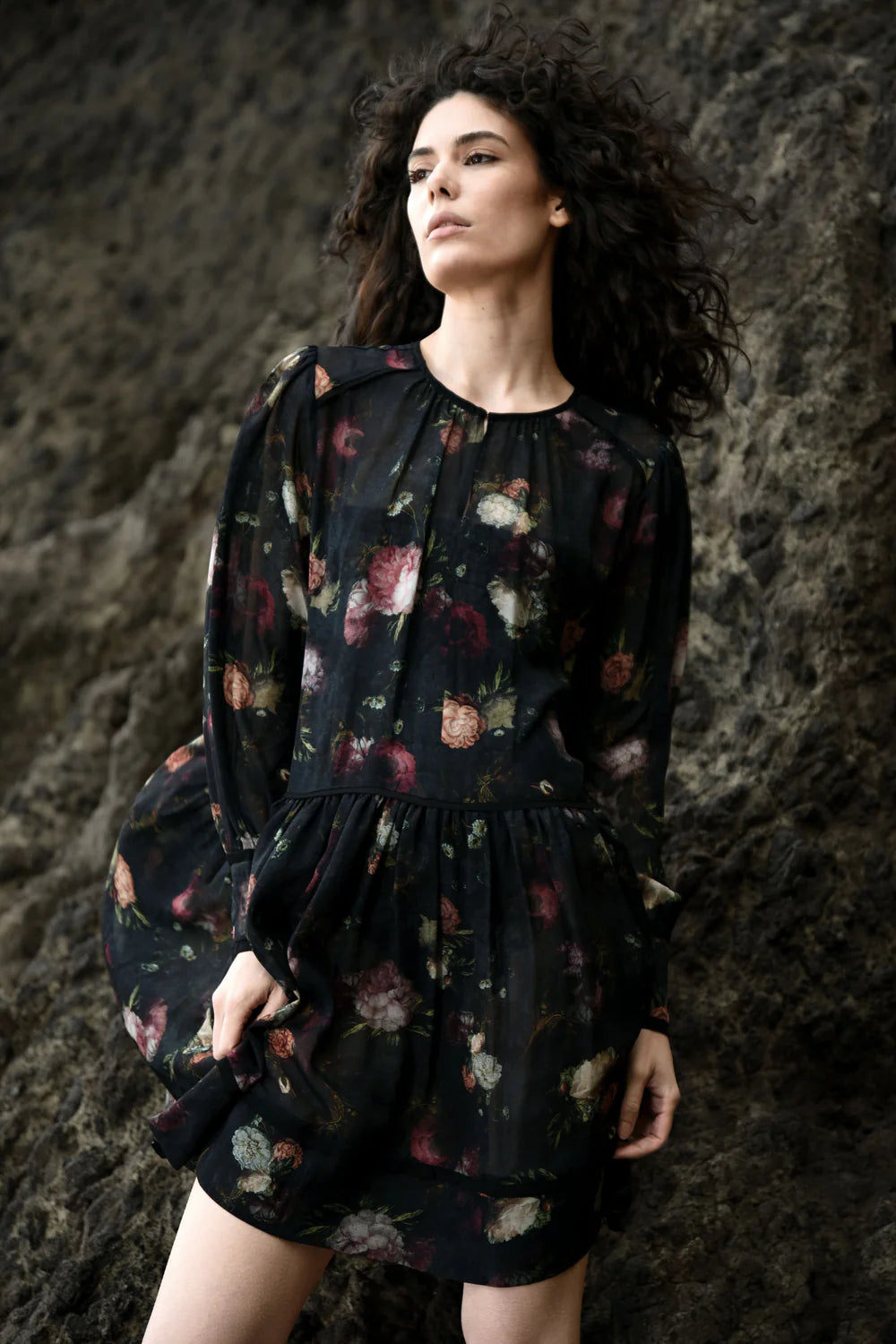 A woman posing on a rocky surface wearing a black floral tunic dress with gathered sleeves and a structured cuff.