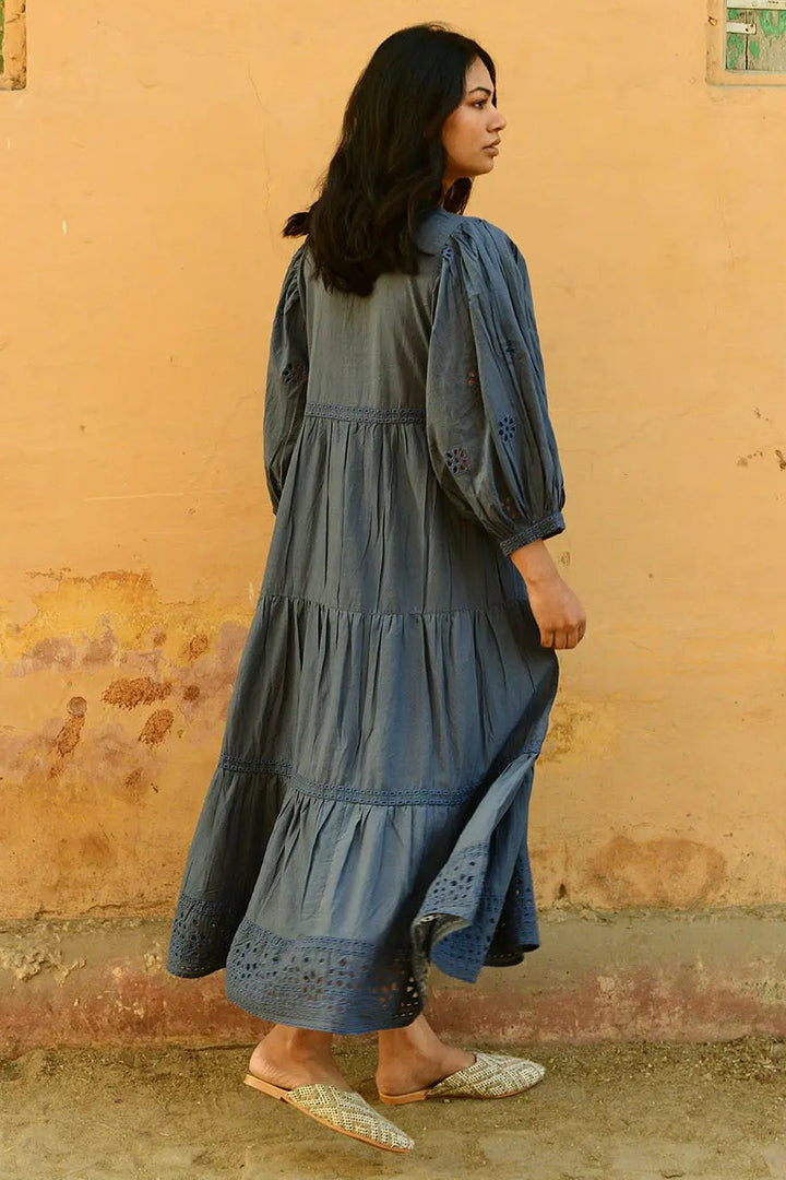 Woman in a long blue dress standing against a yellow wall with green windows.
