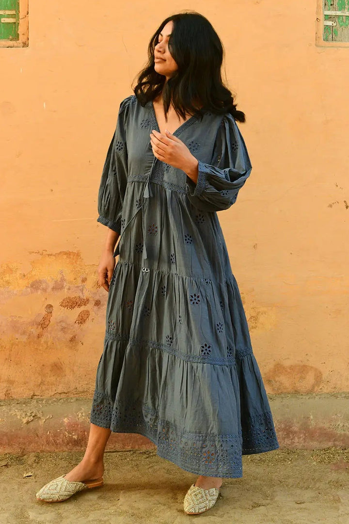 Woman in a long blue dress standing in front of a peach-colored wall with green windows.