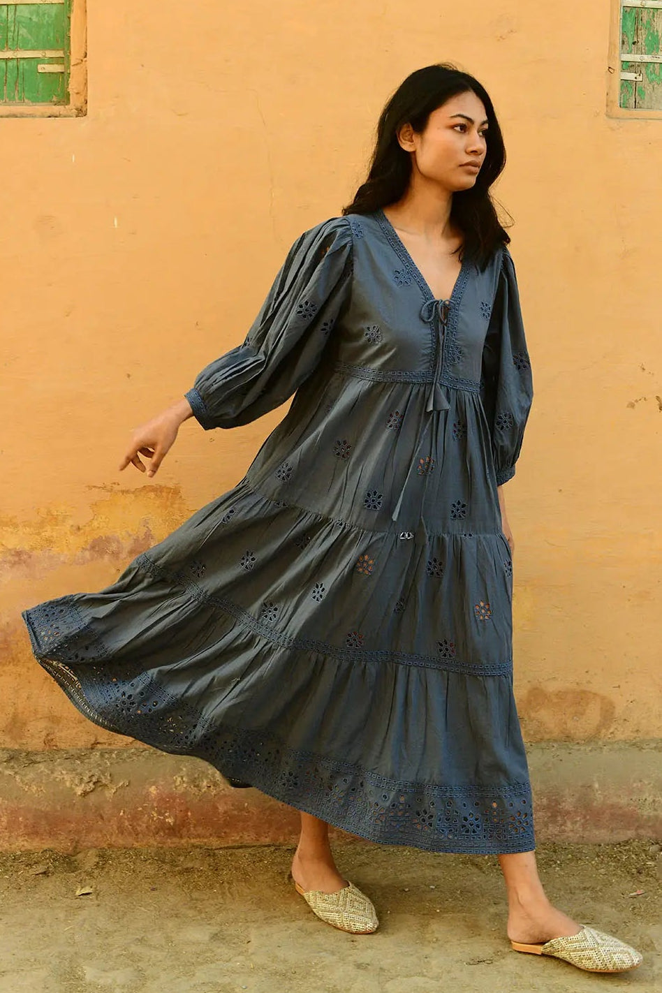 Woman in a blue dress standing against a beige wall with green windows.