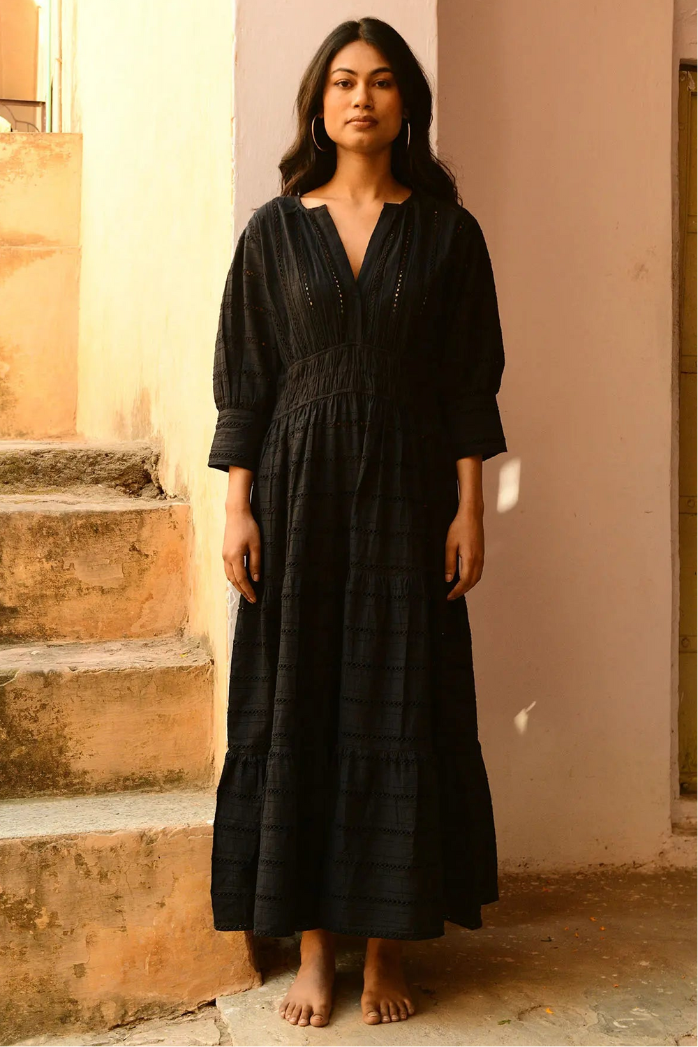 A person standing on a staircase wearing a long black maxi dress with a V-neckline, three-quarter sleeves, and a ruffled hem.