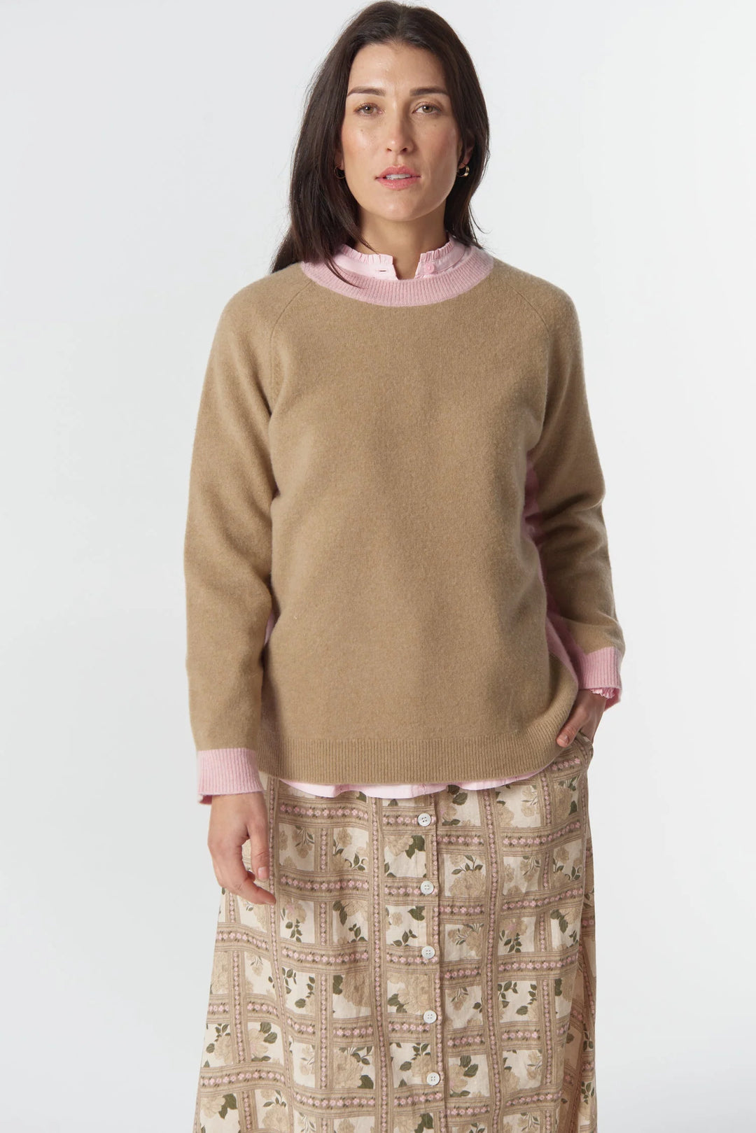 Woman wearing a beige sweater over a pink shirt with a patterned skirt on a white background