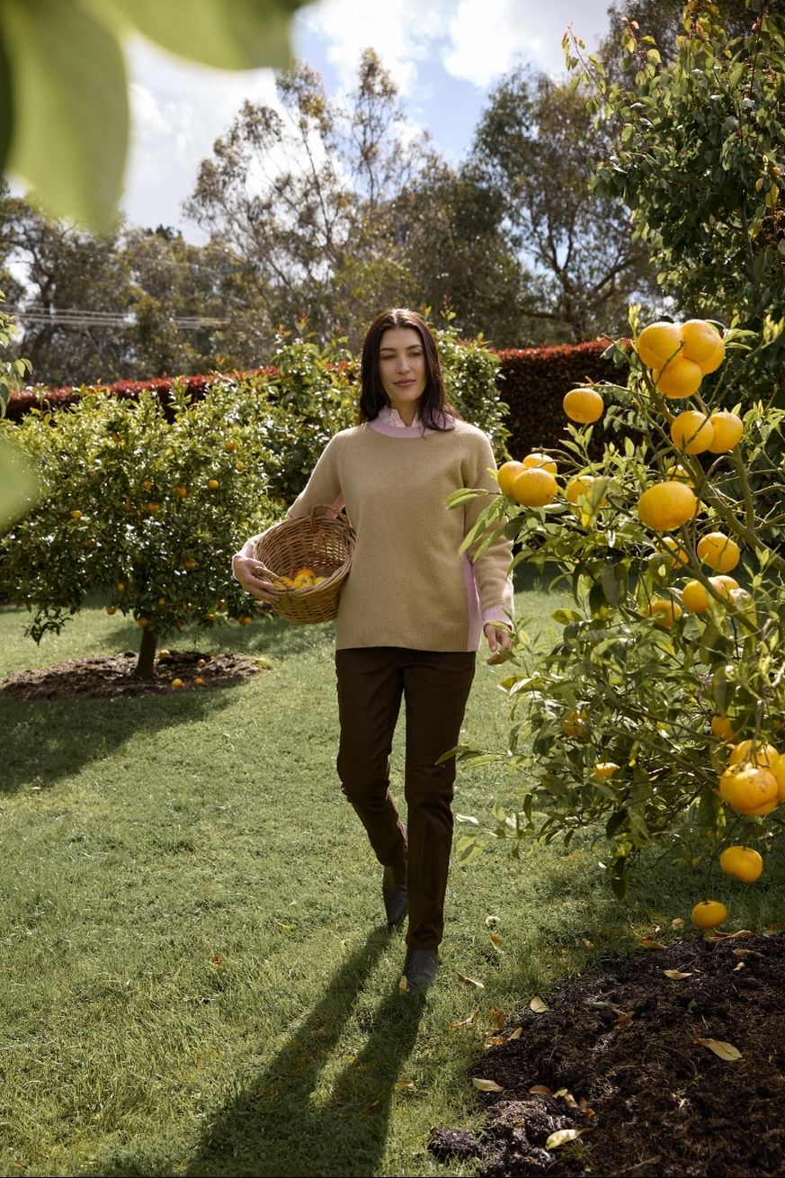 Woman walking through a garden with fruit trees, holding a basket of fruit.