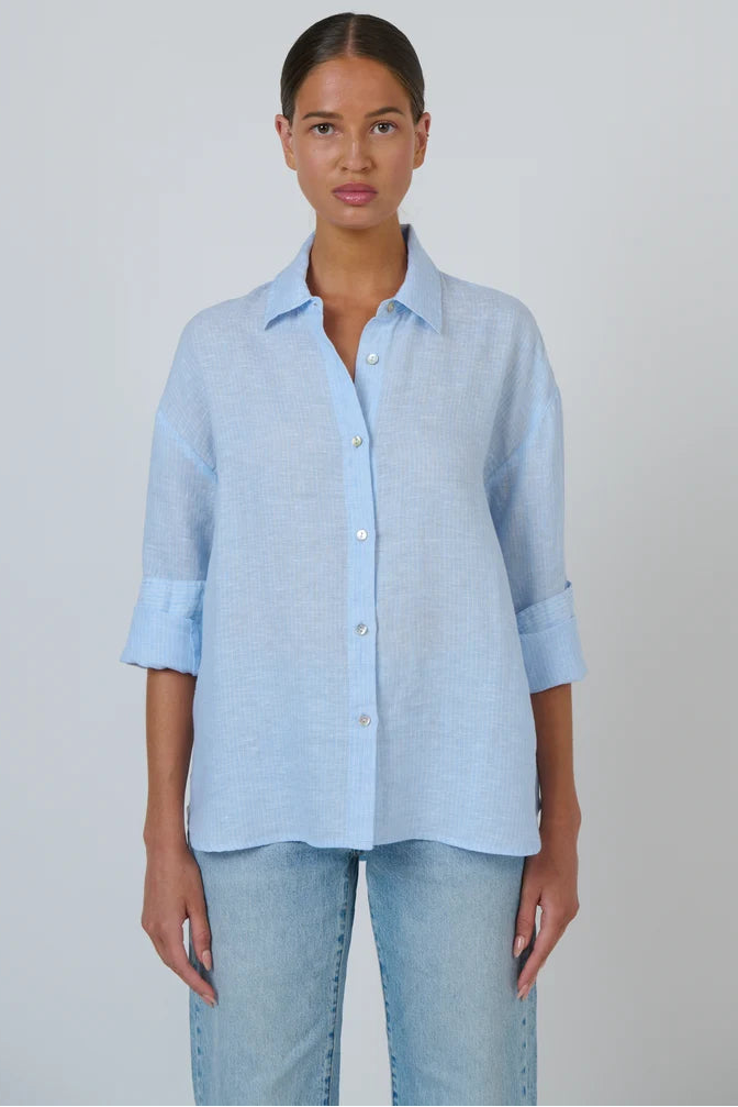 A woman modeling a light blue linen button-up shirt with three-quarter sleeves and light-wash jeans, standing against a neutral light gray background.