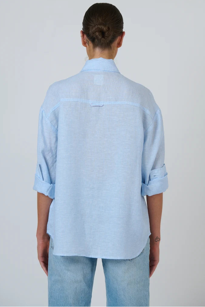 A person shown from the back wearing a light blue linen button-up shirt with rolled sleeves and light blue jeans, photographed against a plain light gray studio background.