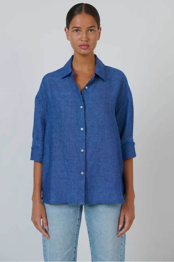 A woman modeling a blue textured button-up shirt with rolled-up sleeves and light-wash jeans, standing against a plain light gray studio background.