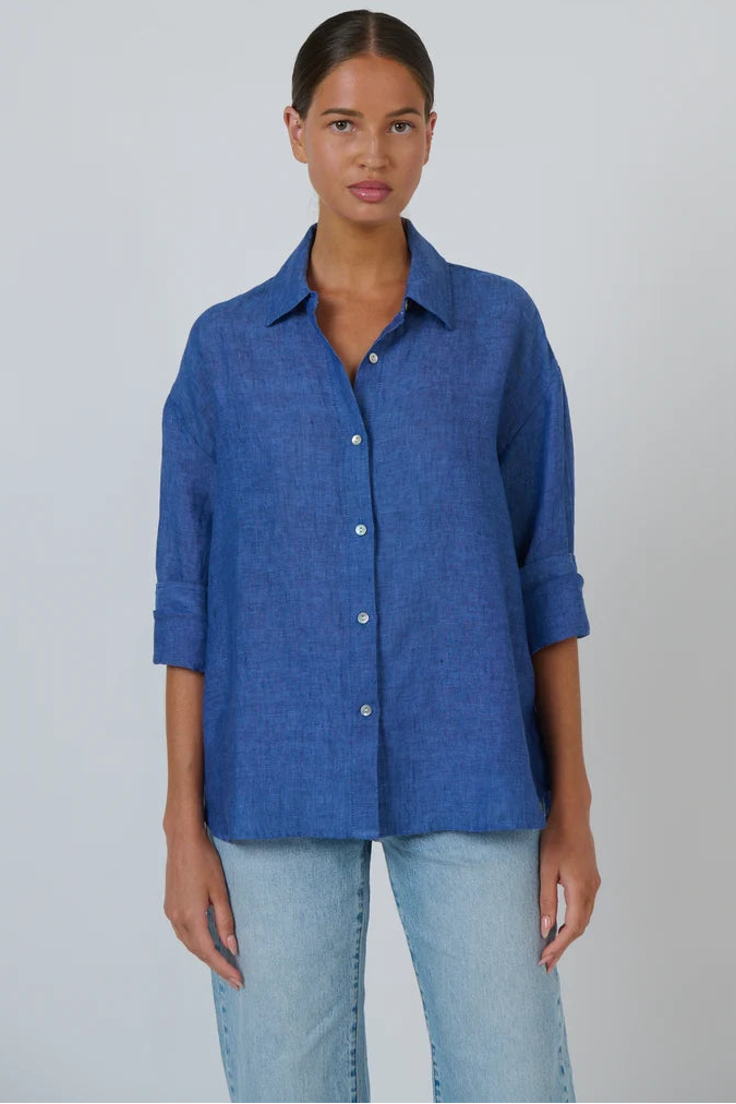 A woman modeling a blue button-up shirt with light-wash jeans against a plain light gray studio background.