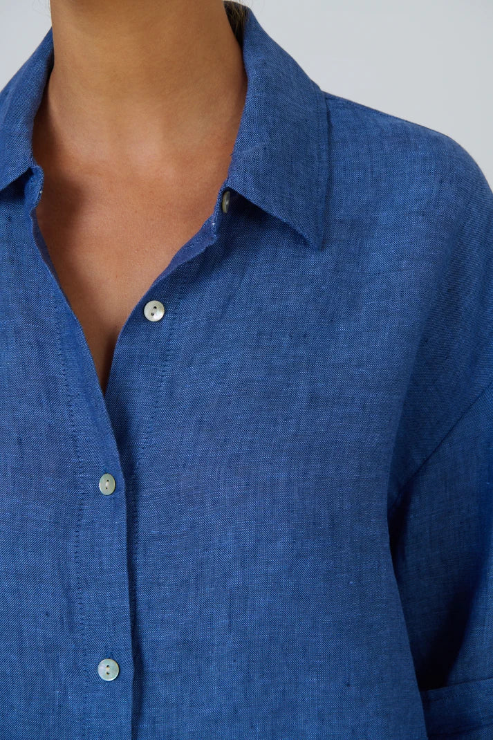 Close-up of a person wearing a blue button-front collared shirt, showing the collar, chest area, and part of the torso against a light neutral background.