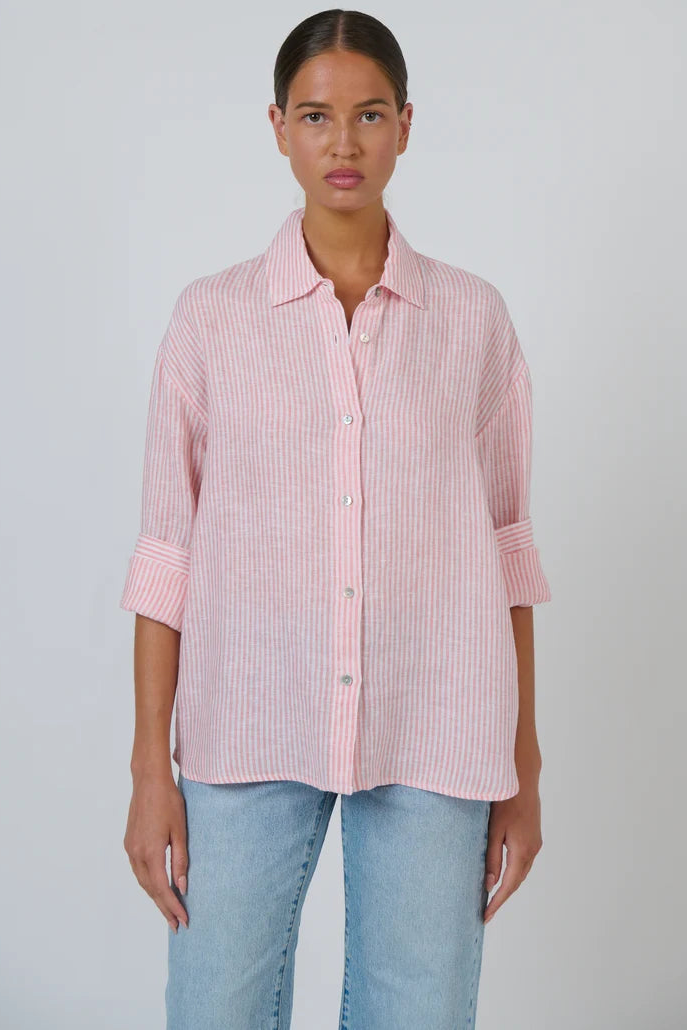 A woman modeling a light pink and white vertically striped button-down shirt with rolled-up sleeves, paired with light blue jeans, against a plain light gray background.