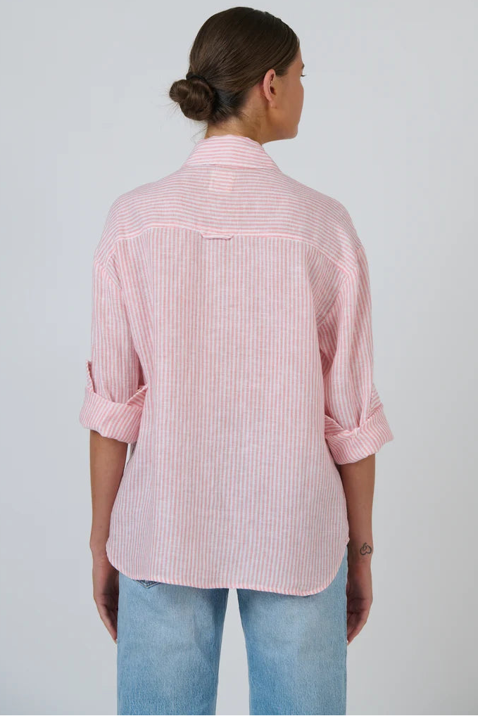 A woman viewed from the back wearing a light pink and white vertically striped button-down shirt with rolled-up sleeves, paired with light blue jeans, against a plain light gray studio background.