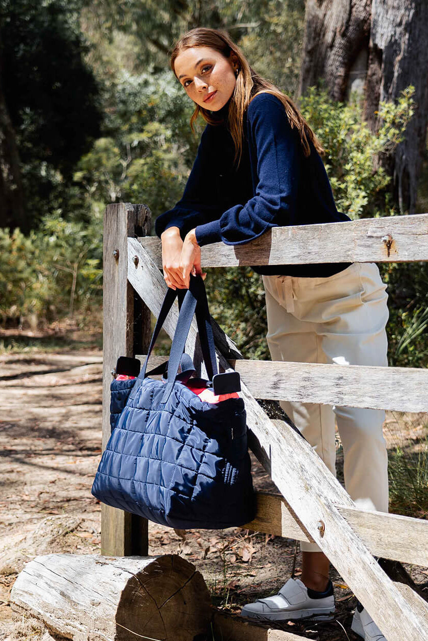 Woman holding a blue quilted bag outdoors near a wooden fence.