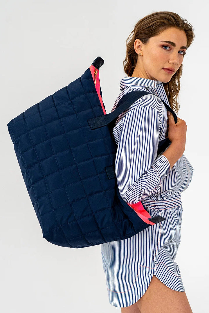 A woman wearing a blue and white striped shirt and shorts carries a large navy blue quilted tote bag with bright pink accents on a plain white studio background.