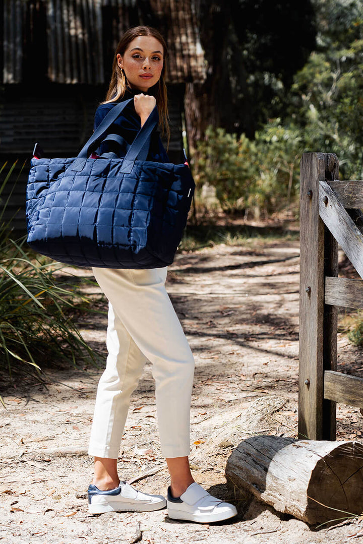 Woman holding a blue quilted bag in a natural setting with a wooden fence and greenery.