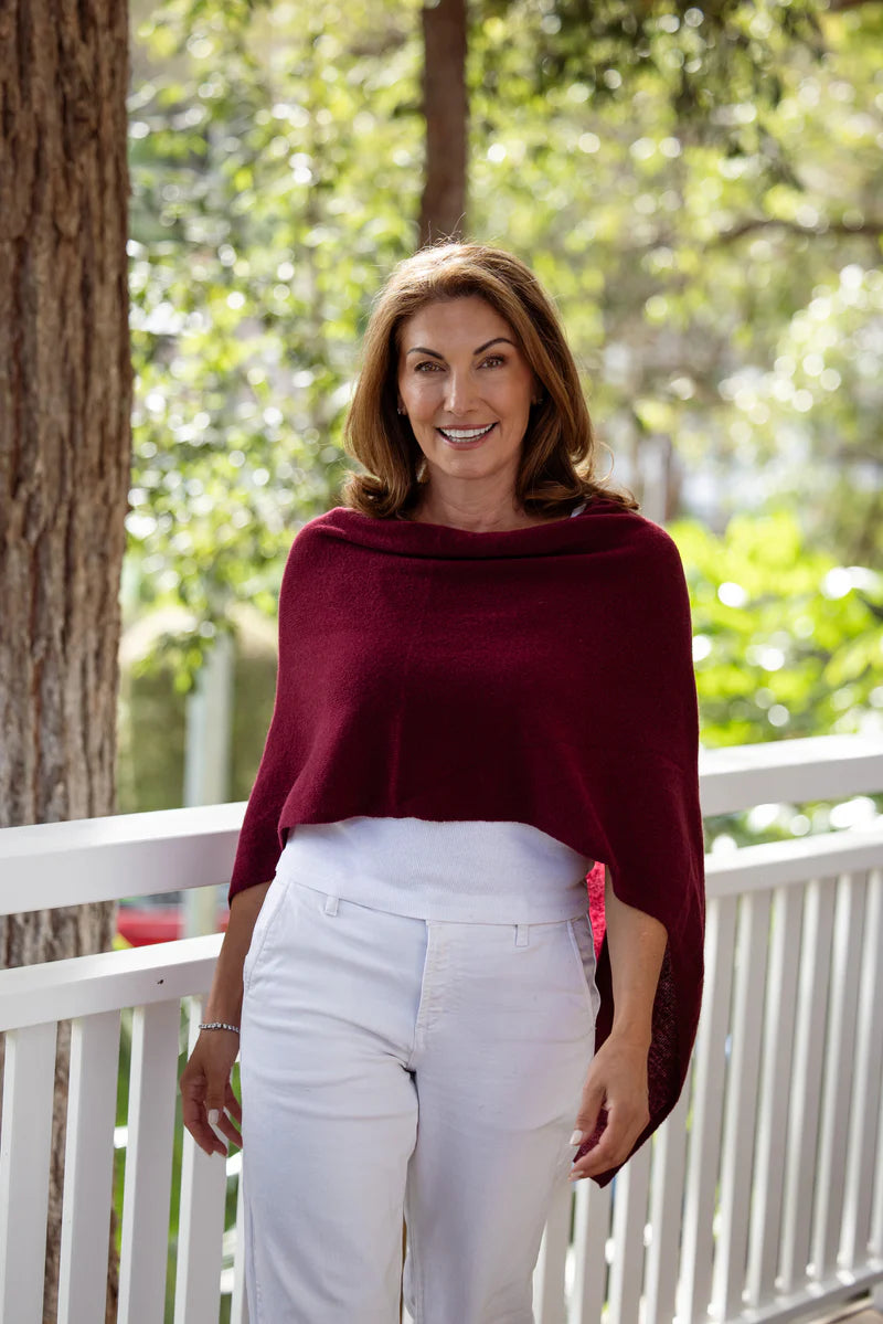 Woman wearing a burgundy sweater and white pants standing on a porch with greenery in the background