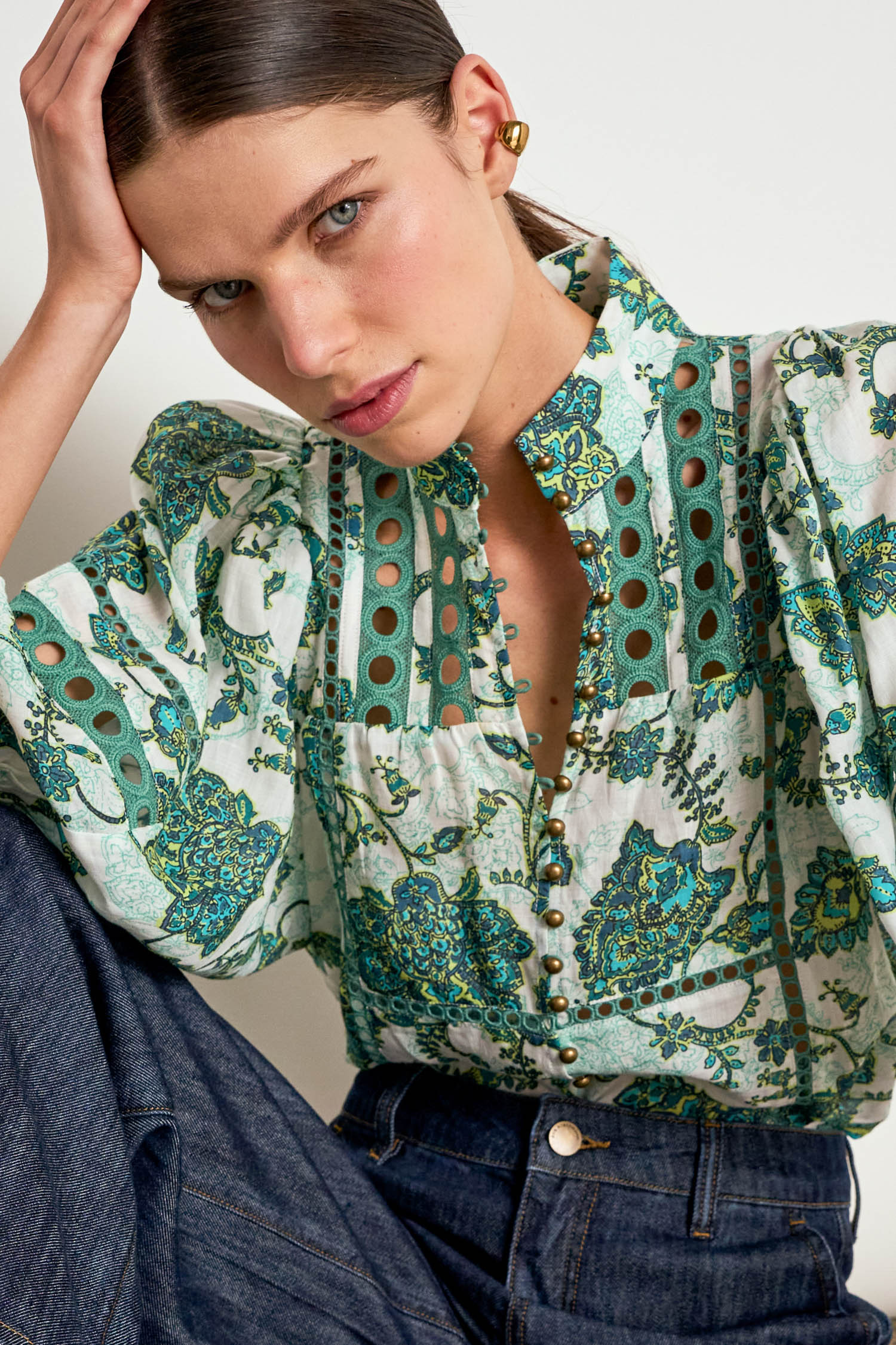 A close-up of a woman wearing a green and teal paisley print blouse with eyelet cutouts and dark blue jeans, posing against a light neutral background.