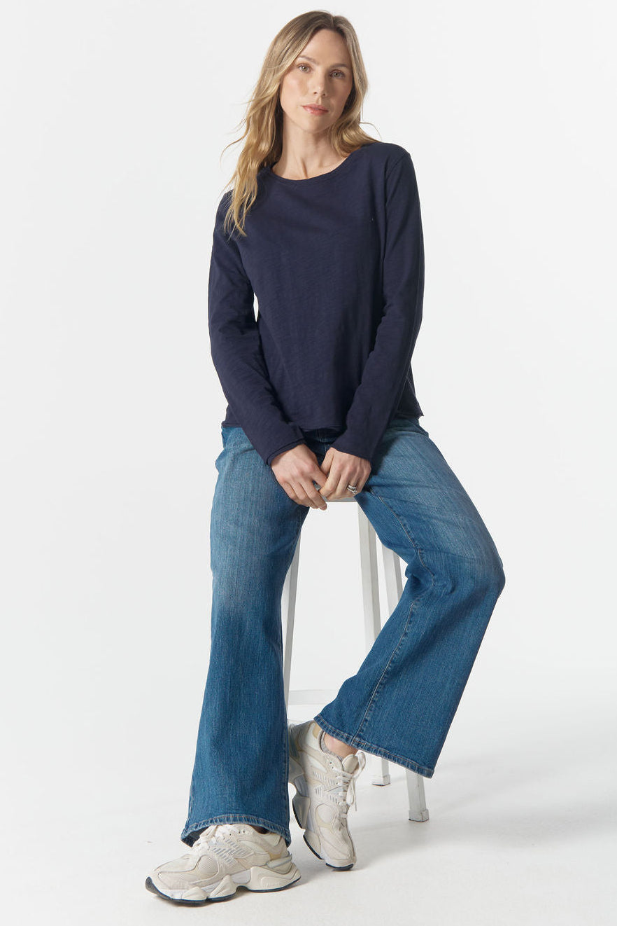 Woman sitting on a stool wearing a navy sweater and blue jeans against a white background