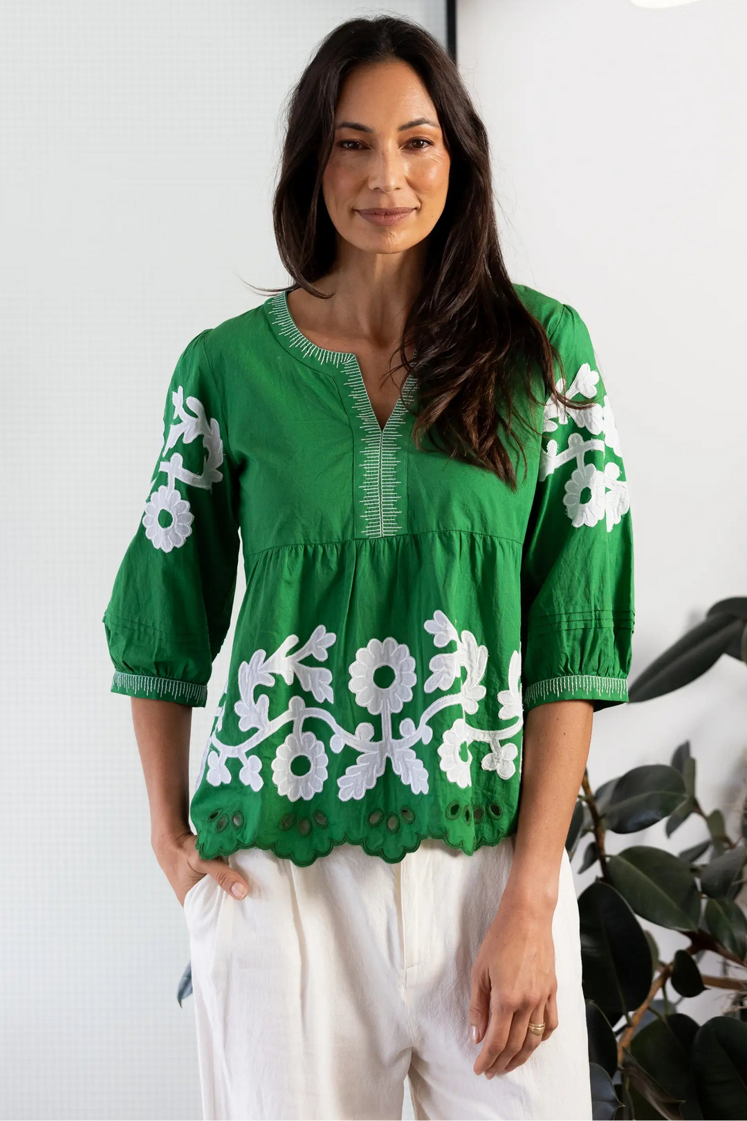 A woman wearing a vibrant green embroidered blouse with white floral patterns and white pants, standing in a modern room with a white wall, black horizontal bar, and a floor lamp.