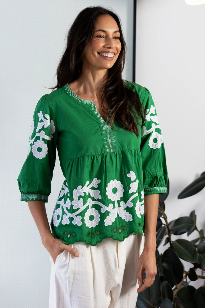 A woman wearing a vibrant green embroidered blouse with white floral patterns and white pants, standing in a modern room with a black-framed window, a floor lamp, and potted plants.