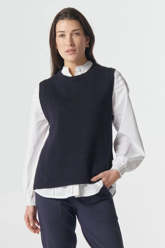 Woman wearing a navy sleeveless top, white shirt, and dark pants on a white background