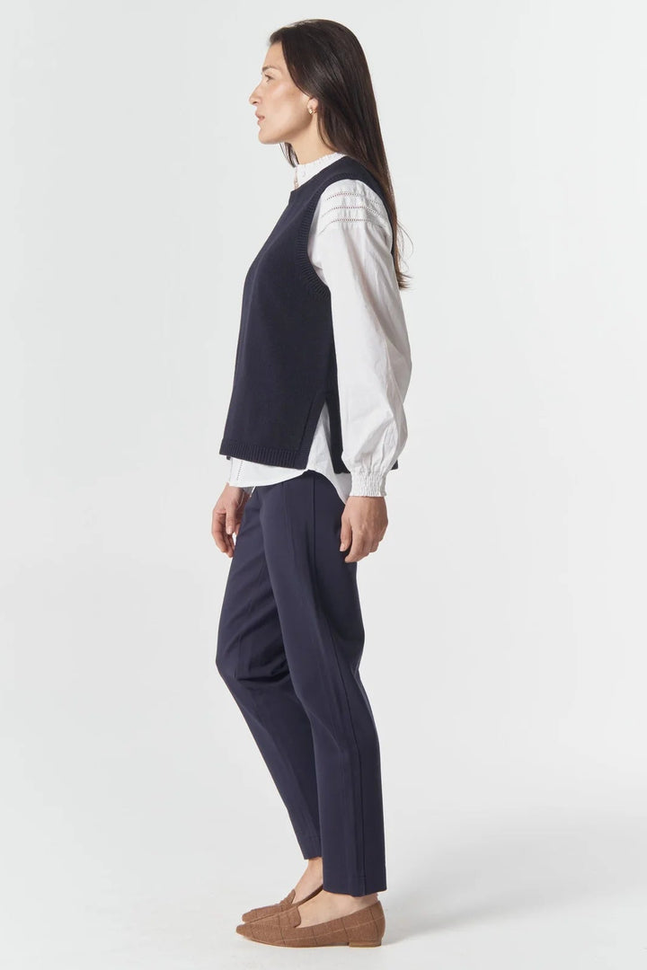 Woman wearing a navy vest over a white shirt with dark pants on a white background