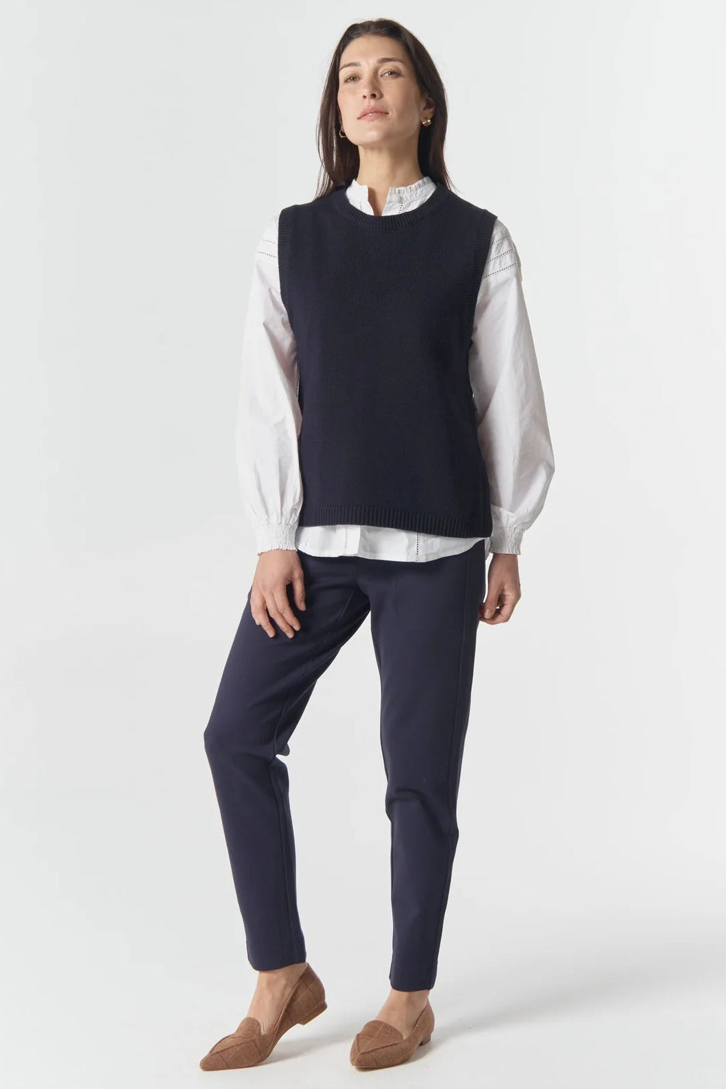 Woman wearing a navy sleeveless sweater over a white shirt with dark pants on a white background