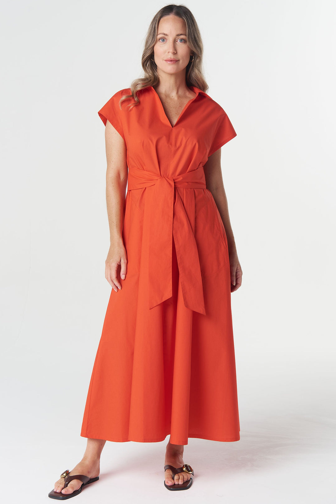 A woman modeling a bright orange wrap-style midi dress with short sleeves, a V-neckline, and a self-tie waist, paired with dark brown sandals against a plain light gray studio background.