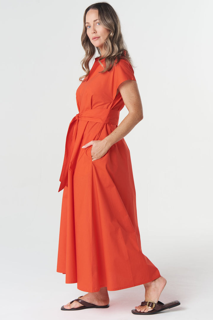 A woman modeling a bright orange-red wrap-style maxi dress with short sleeves, a self-tie waist belt, and side pockets, paired with black sandals, against a plain light gray studio background.