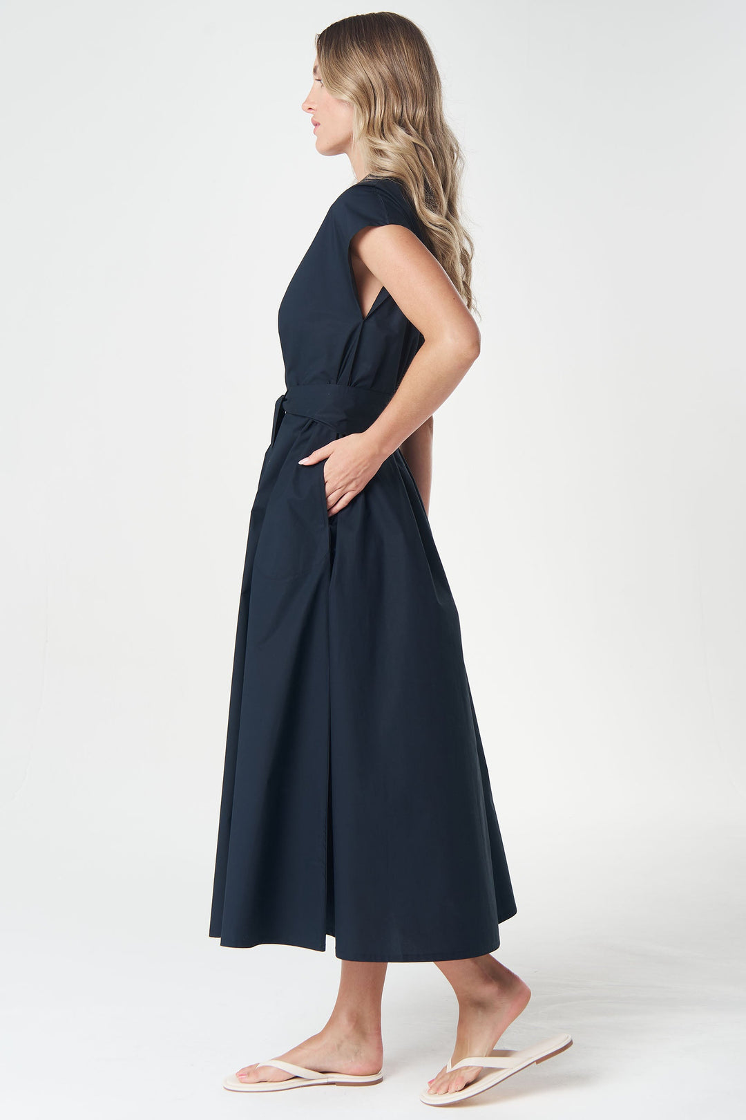 A woman modeling a sleeveless navy midi dress with a side slit and belt, shown from the side against a plain white studio background.