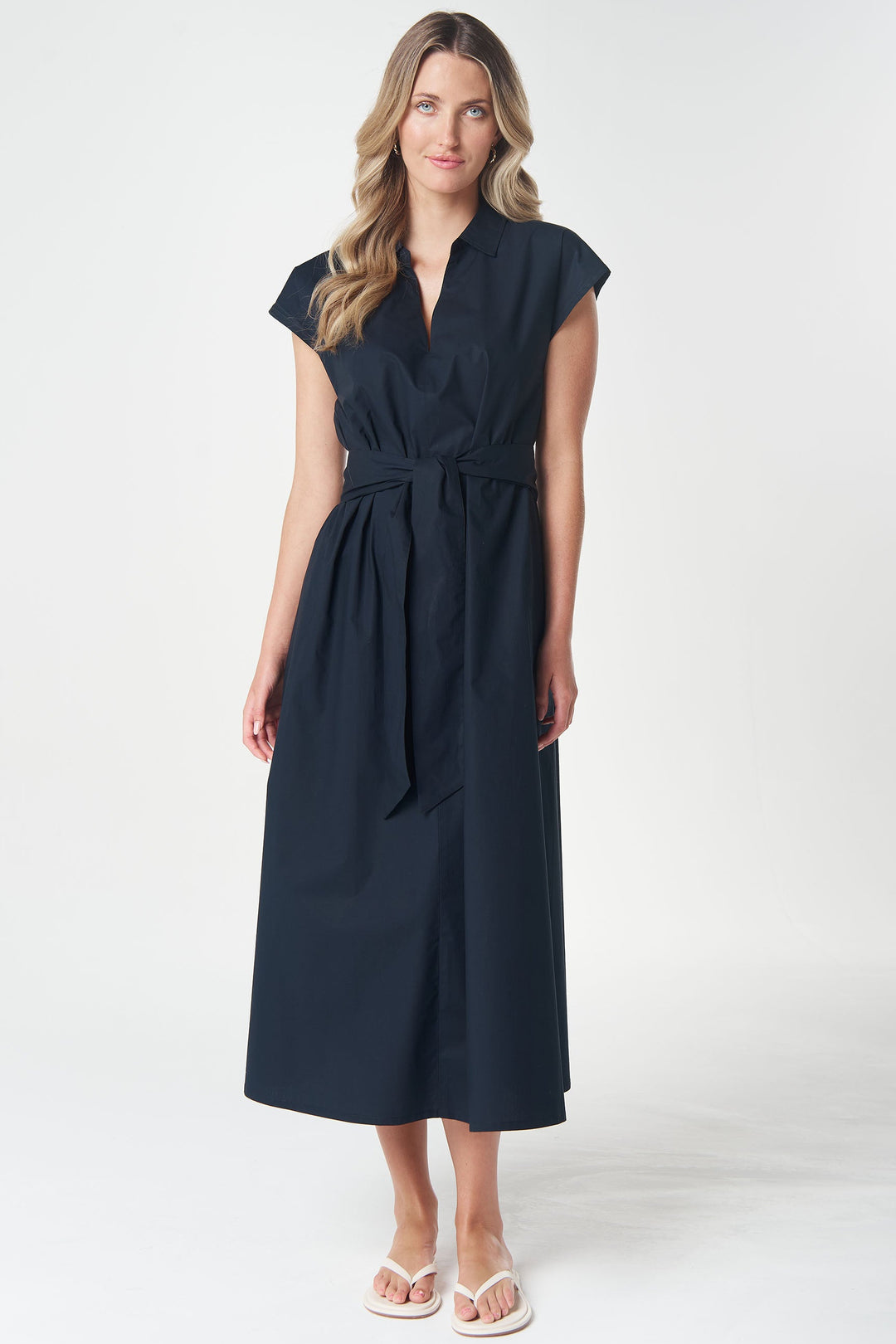 A woman modeling a dark navy midi dress with a wrap-style V-neck, cap sleeves, a matching fabric tie belt at the waist, and a straight skirt, paired with white flip-flop sandals against a light gray studio background.